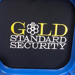 Gold Standard Security logo embroidered in gold and white thread on a black garment, still in the embroidery hoop at Dad Bod Screen Printing